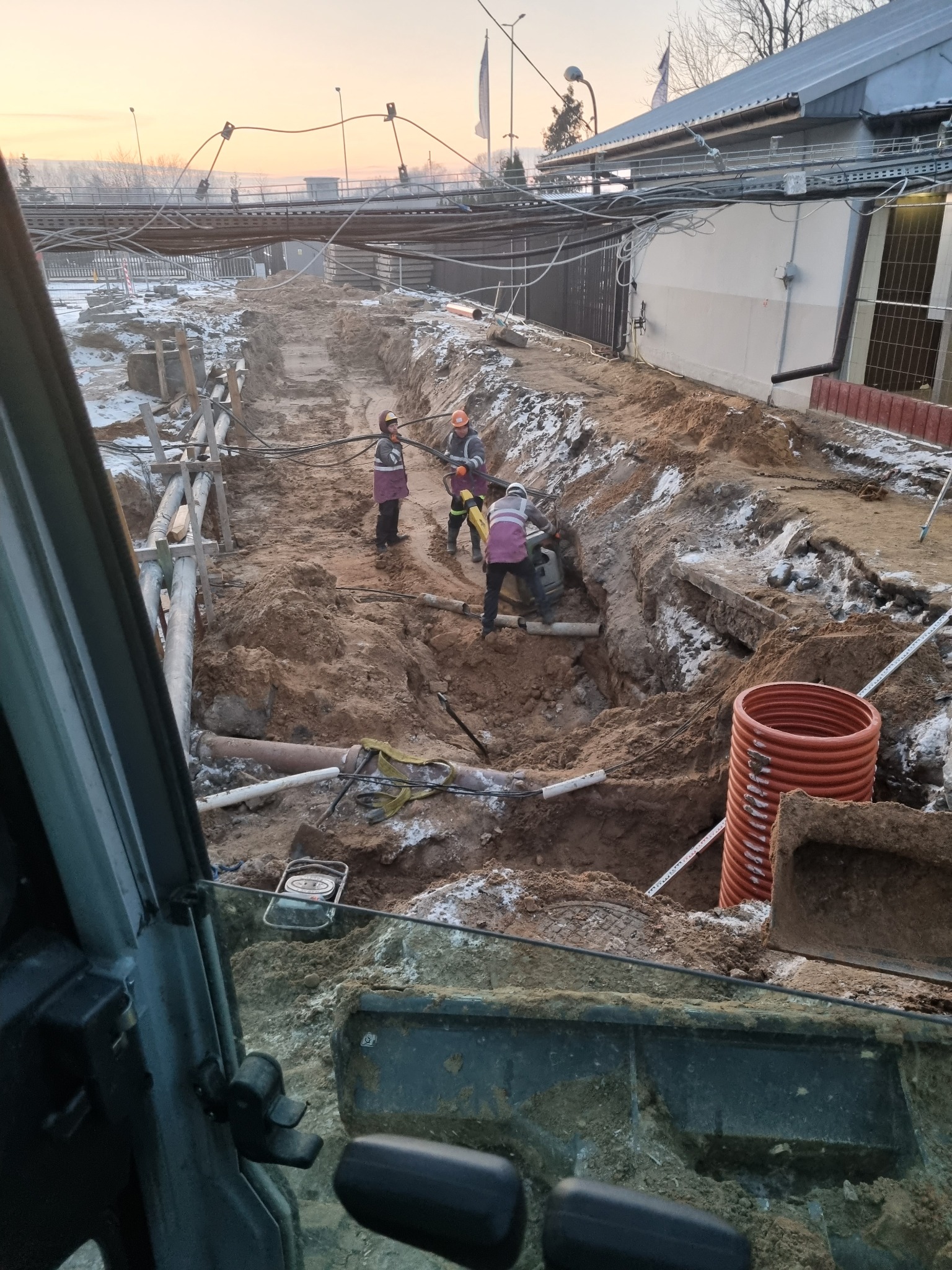 Prace ziemne przy budowie przyłącza wodno-kanalizacyjnego; widoczne rury, wykop, pracownicy w kaskach i odblaskowych kamizelkach, częściowo ośnieżona ziemia, ujęcie z wnętrza kabiny maszyny...