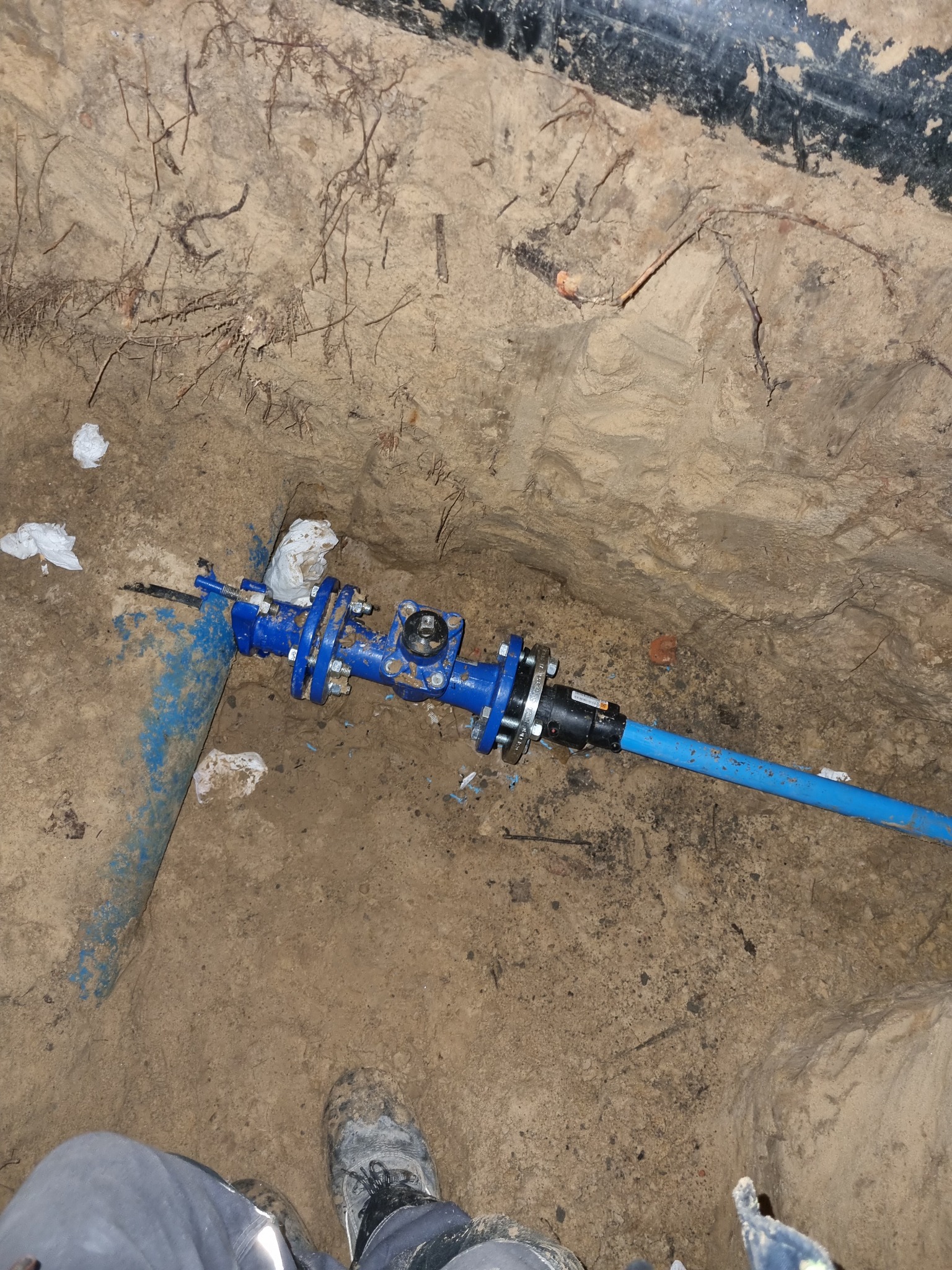 Niebieskie rury wodociągowe połączone z zaworem w wykopie, widoczne ślady ziemi i wilgoci, fragment buta roboczego w prawym dolnym rogu.
