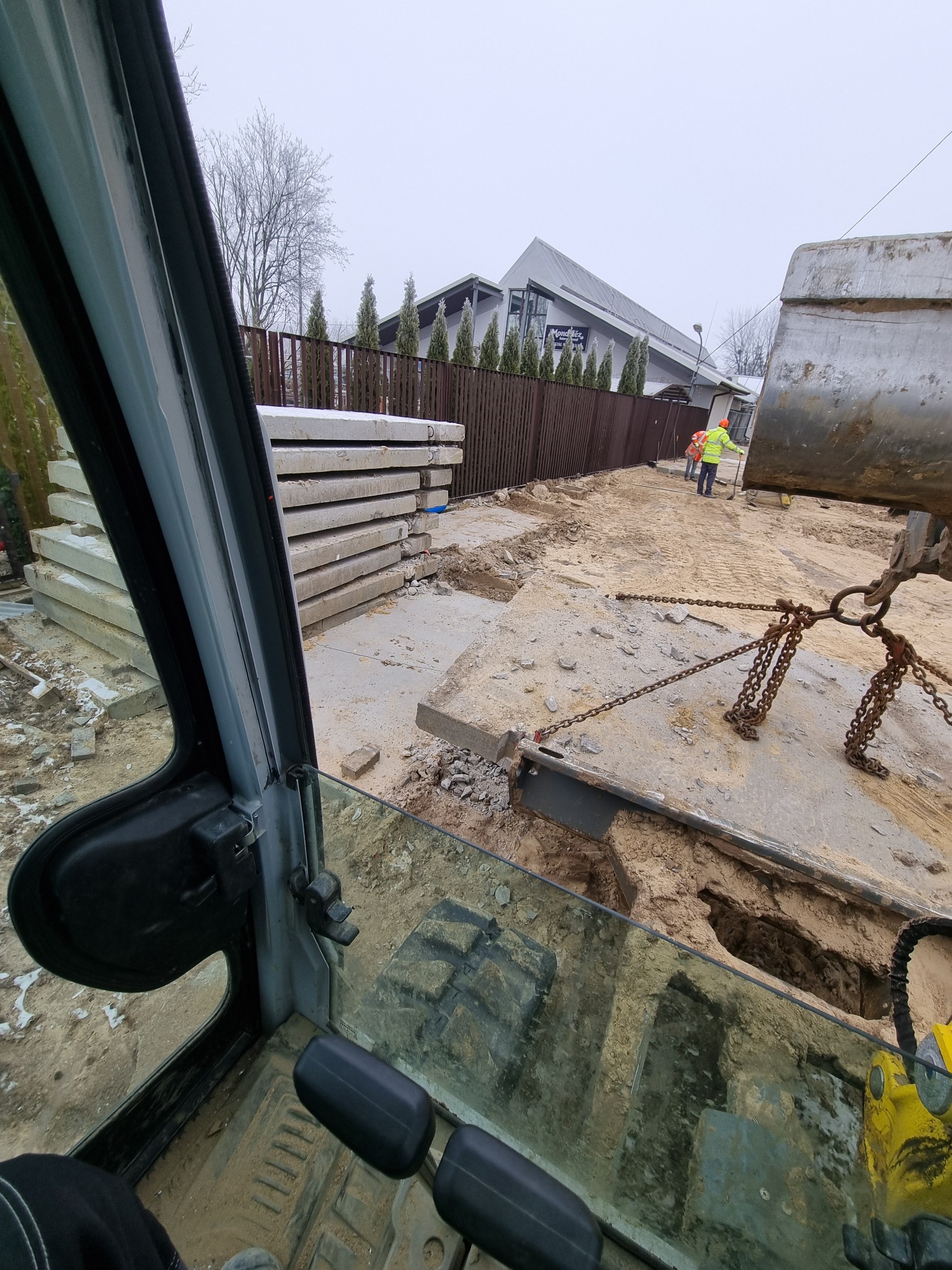 Widok z kabiny koparki na plac budowy z betonowymi płytami i pracownikami w odblaskowych ubraniach, przygotowywany wykop pod przyłącze wodno-kanalizacyjne