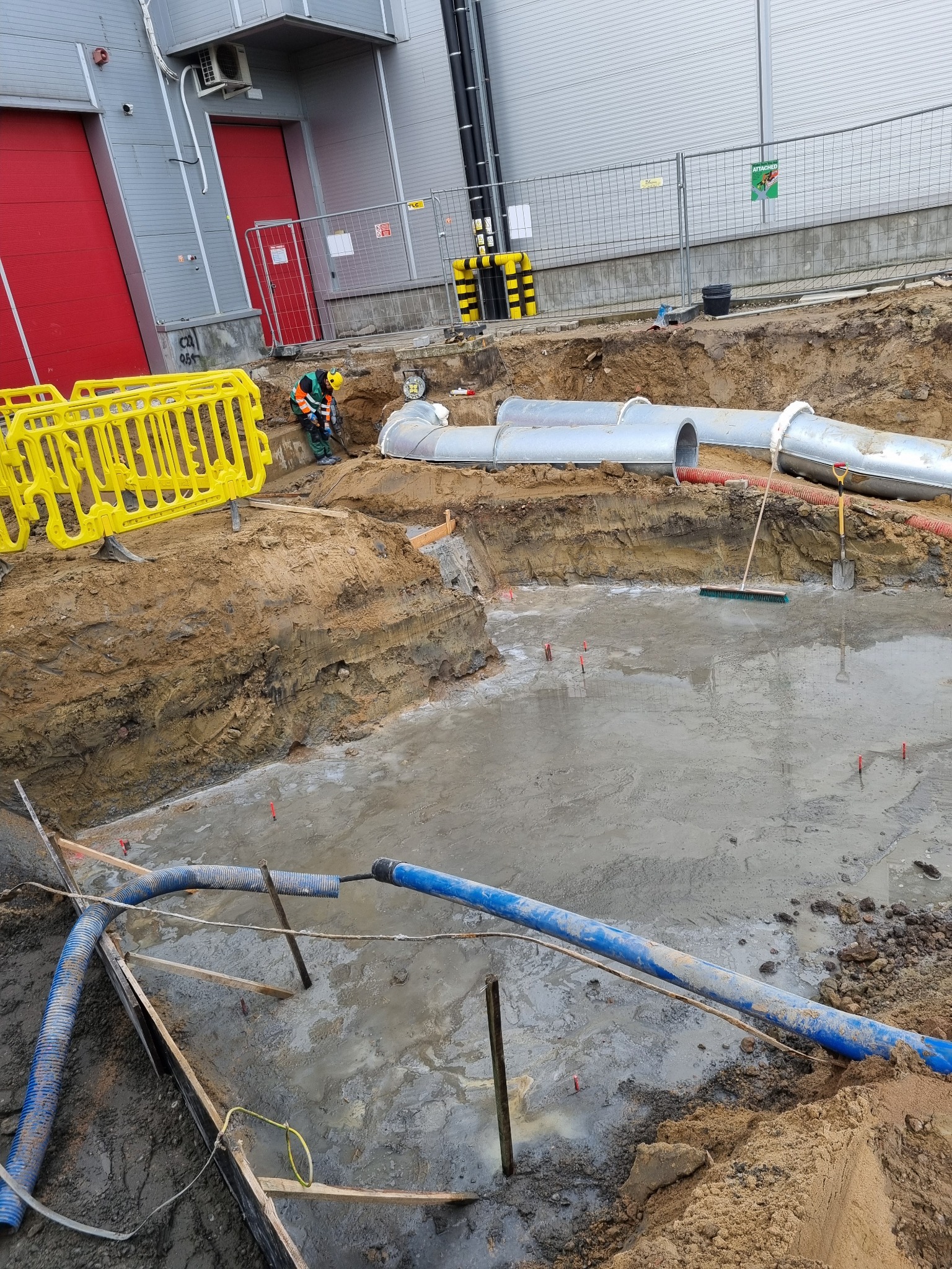 Wykop z częściowo zalaną wodą, widoczne rury kanalizacyjne, pracownik w odblaskowej kamizelce i kasku przy wykopie, żółte barierki zabezpieczające oraz budynek przemysłowy w tle.