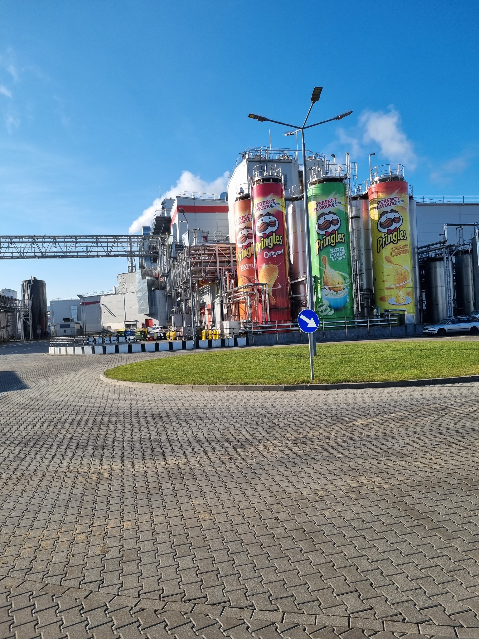 Widok na fabrykę z dużymi cylindrycznymi zbiornikami z reklamami Pringles, kominy z lekkim dymem na tle błękitnego nieba, kostka brukowa na pierwszym planie.