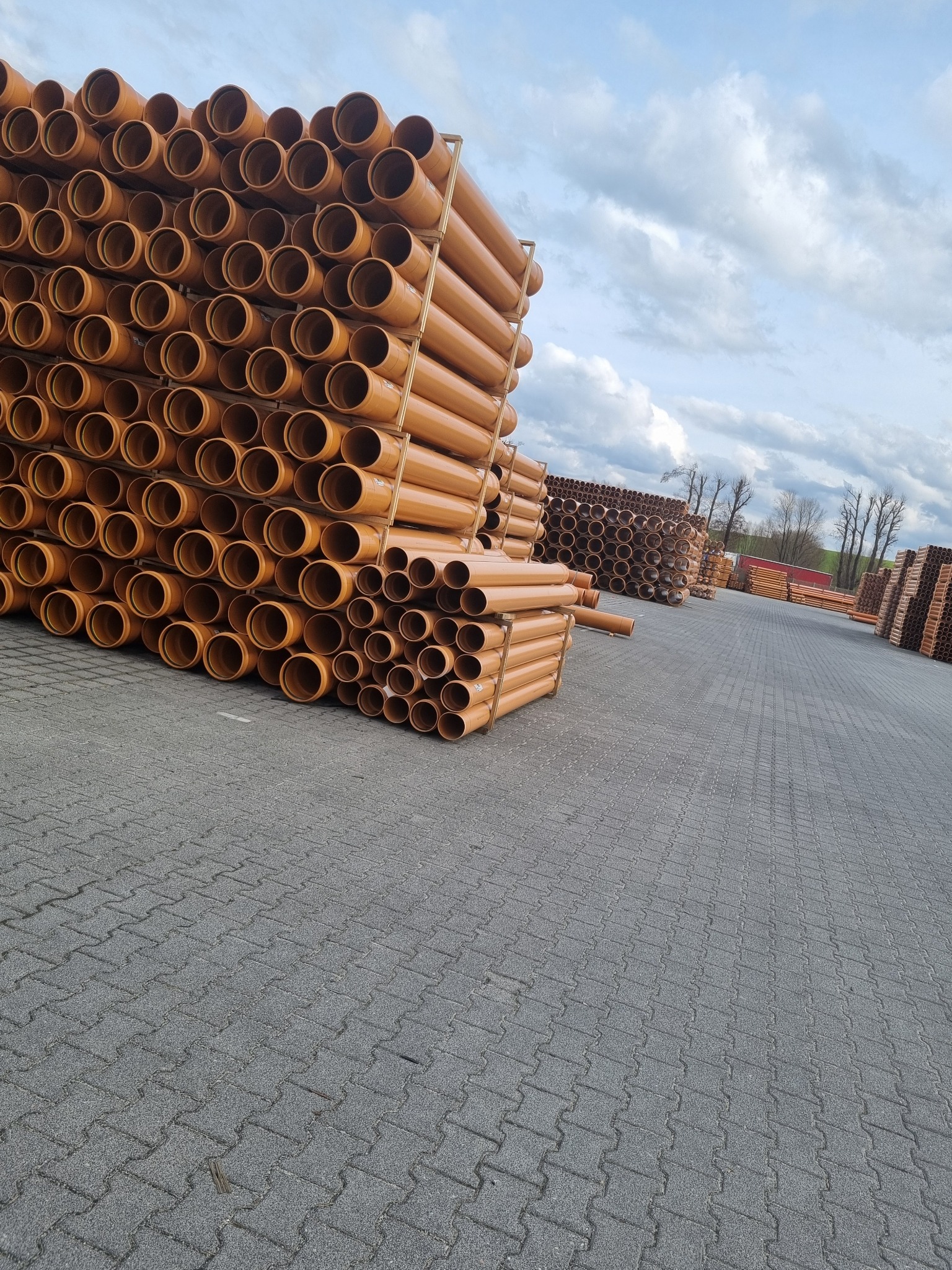Stosy rur kanalizacyjnych z tworzywa sztucznego w kolorze terakoty, ułożone na paletach na placu z kostki brukowej, w tle inne stosy i błękitne niebo z chmurami.