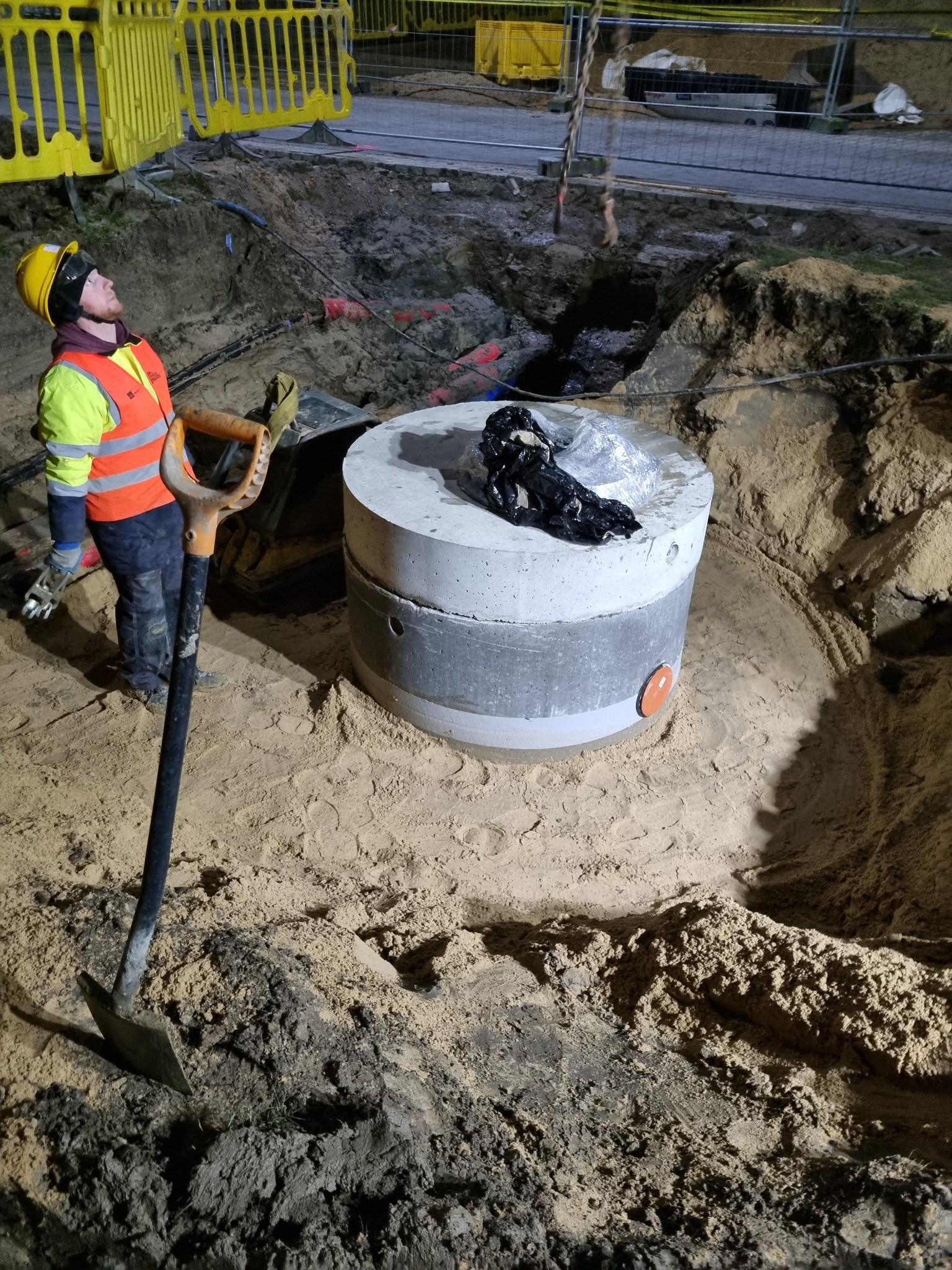 Studzienka kanalizacyjna betonowa w wykopie, pracownik w odblaskowej kamizelce i kasku spogląda w górę, obok łopata wbita w ziemię, teren budowy zabezpieczony barierami.