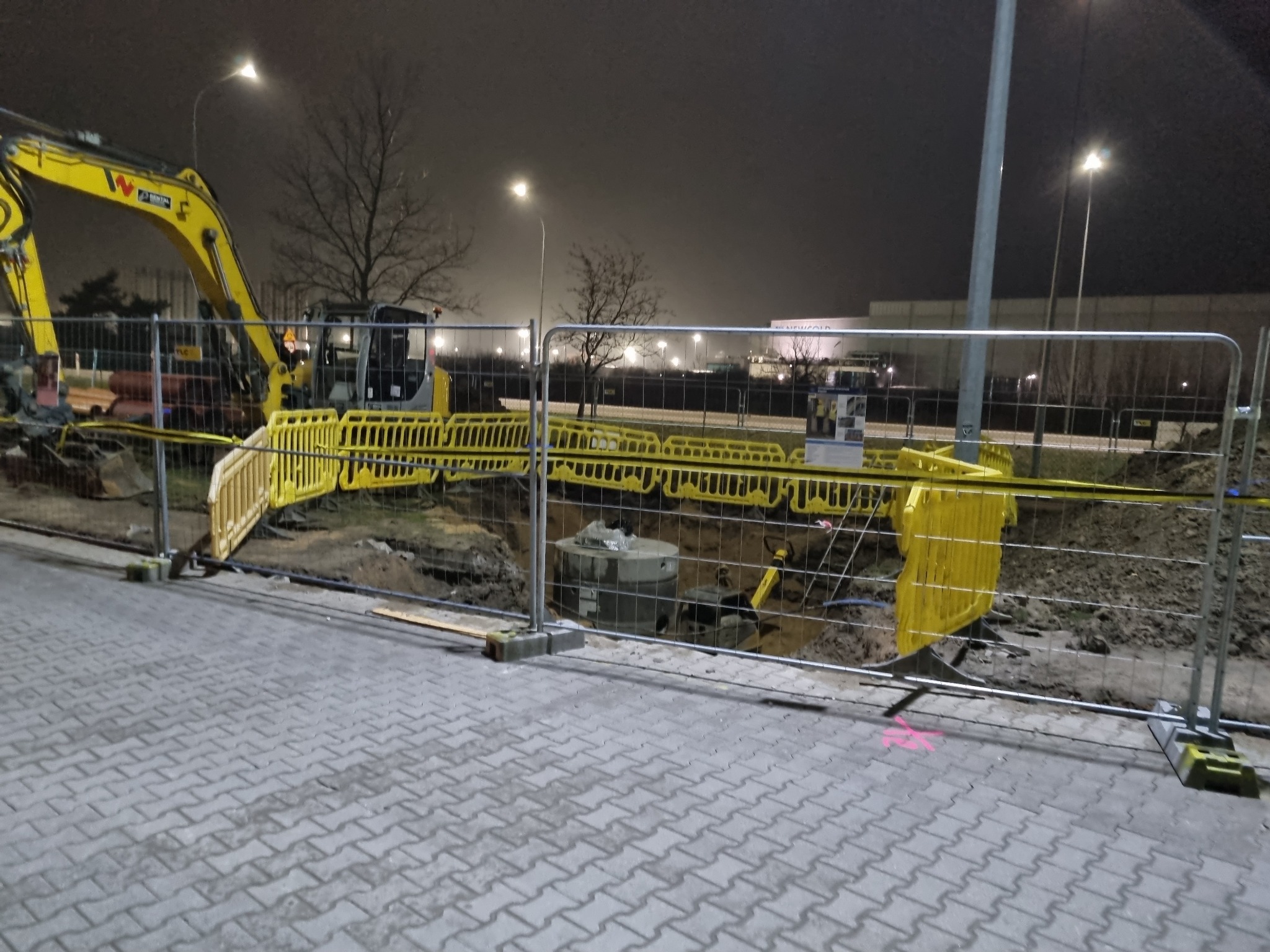 Wykop zabezpieczony żółtą barierką, widoczny fragment koparki i betonowy krąg w ziemi, oświetlenie uliczne w tle, nocna sceneria.
