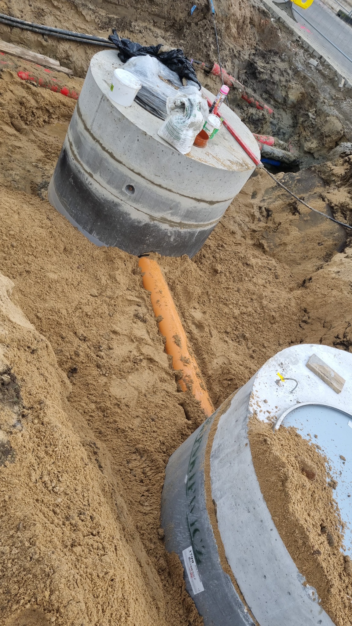 Wykonany wykop z widocznymi betonowymi elementami studni kanalizacyjnej i pomarańczową rurą, zasypane piaskiem. Na górnym elemencie studni leżą narzędzia i materiały budowlane.