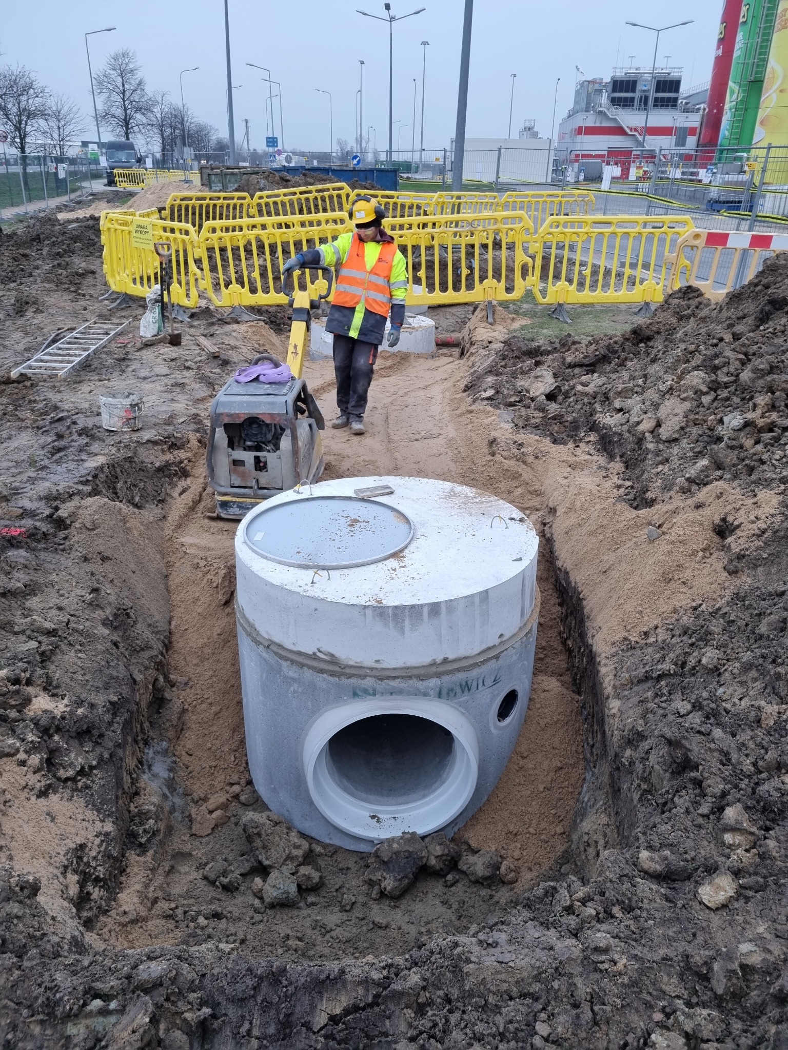 Montaż betonowego zbiornika kanalizacyjnego w wykopie, pracownik zagęszcza grunt ubijarką wibracyjną, zabezpieczenie żółtymi barierkami.
