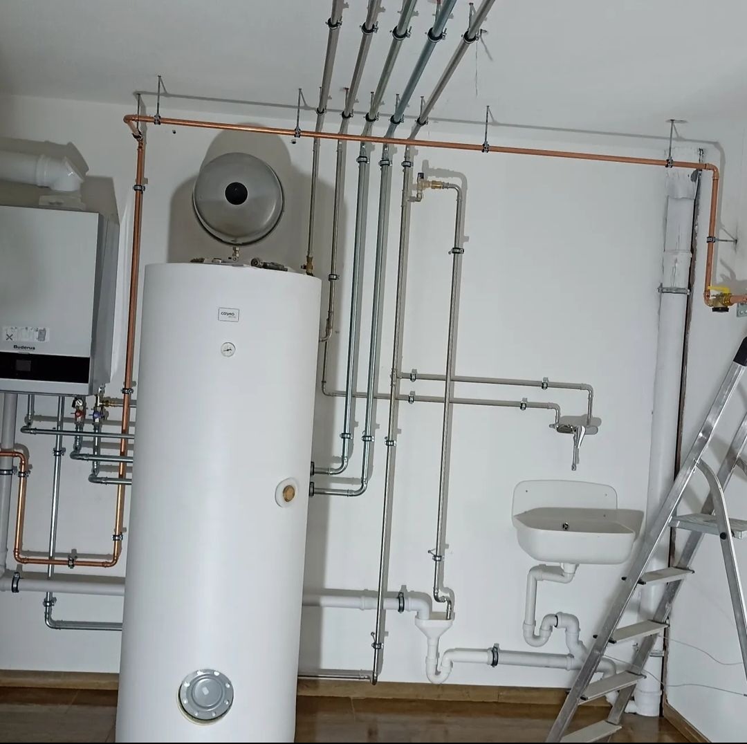 Nowoczesna kotłownia z białym zasobnikiem ciepłej wody, kotłem gazowym Buderus, miedzianymi i stalowymi rurami, umywalką i rozstawioną drabiną aluminiową.