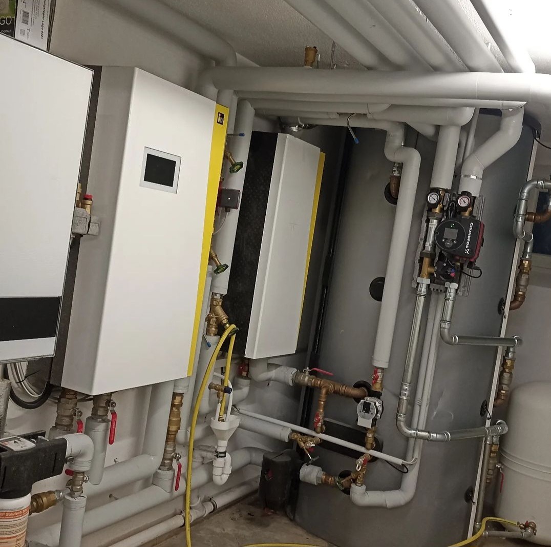 Złożona instalacja grzewcza z dwoma piecami, zbiornikiem akumulacyjnym i licznymi rurami, widok z dołu na szarą ścianę i podłogę.