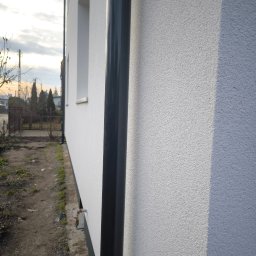 JANEK BUDOWLANIEC - Biała elewacja domu z czarną rurą spustową, widok z boku, z fragmentem ogrodu i niebem w tle. Tekstura tynku barankowego.