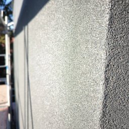 JANEK BUDOWLANIEC - Szara, ziarnista faktura tynku elewacyjnego w zbliżeniu, z widocznym cieniem i fragmentem rusztowania w tle. Delikatne refleksy światła podkreślają strukturę.