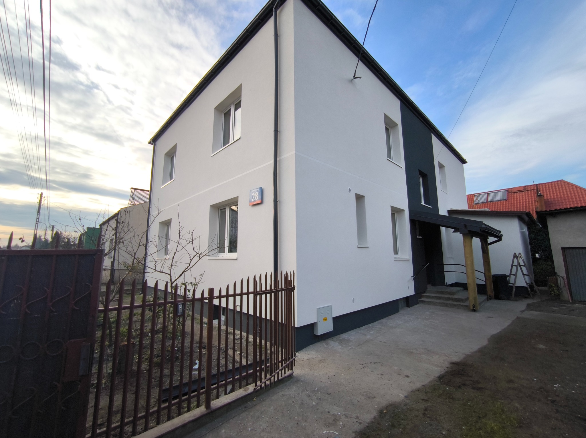 Dwukondygnacyjny dom po renowacji elewacji, z białymi ścianami i antracytowymi akcentami, widoczny zza metalowego ogrodzenia. Na ścianie numer posesji.