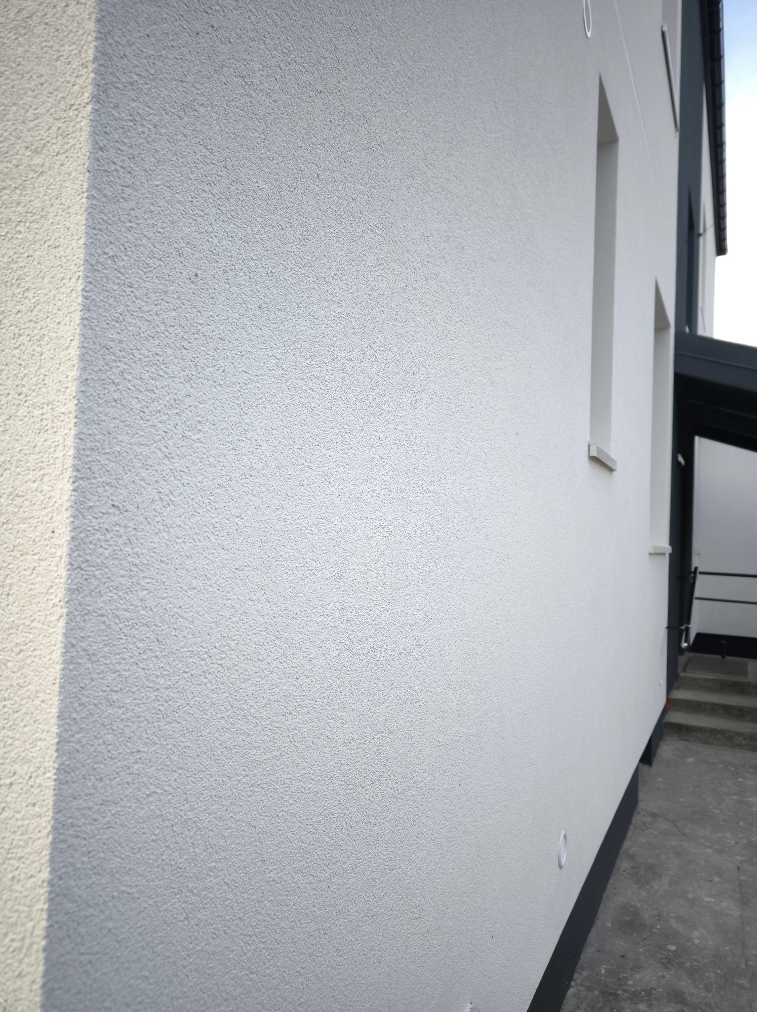 Boczny widok na fragment białej, otynkowanej elewacji domu z widocznymi oknami i wejściem. Tekstura tynku jest wyraźna, a całość utrzymana w minimalistycznym stylu.