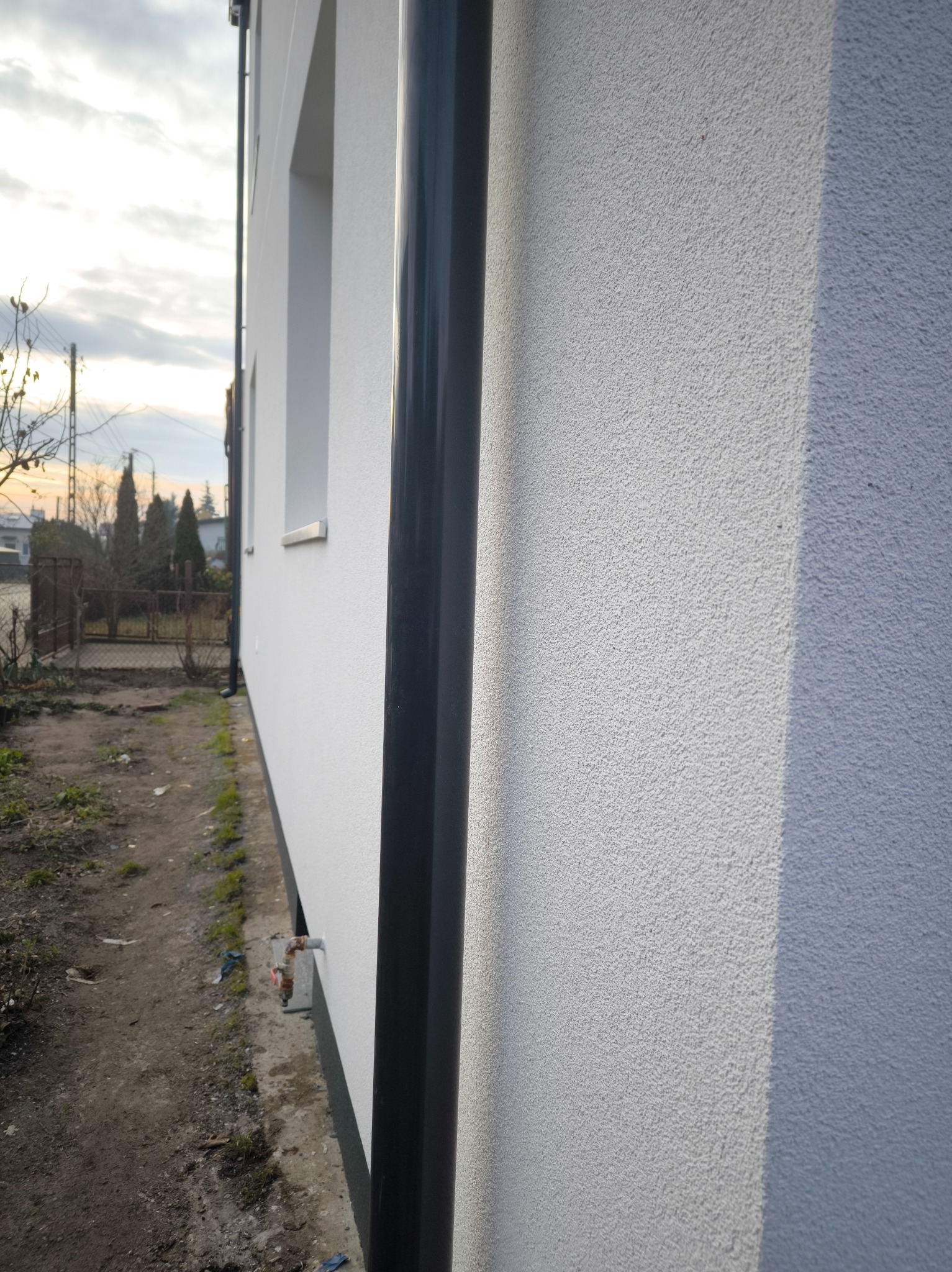 Biała elewacja domu z czarną rurą spustową, widok z boku, z fragmentem ogrodu i niebem w tle. Tekstura tynku barankowego.