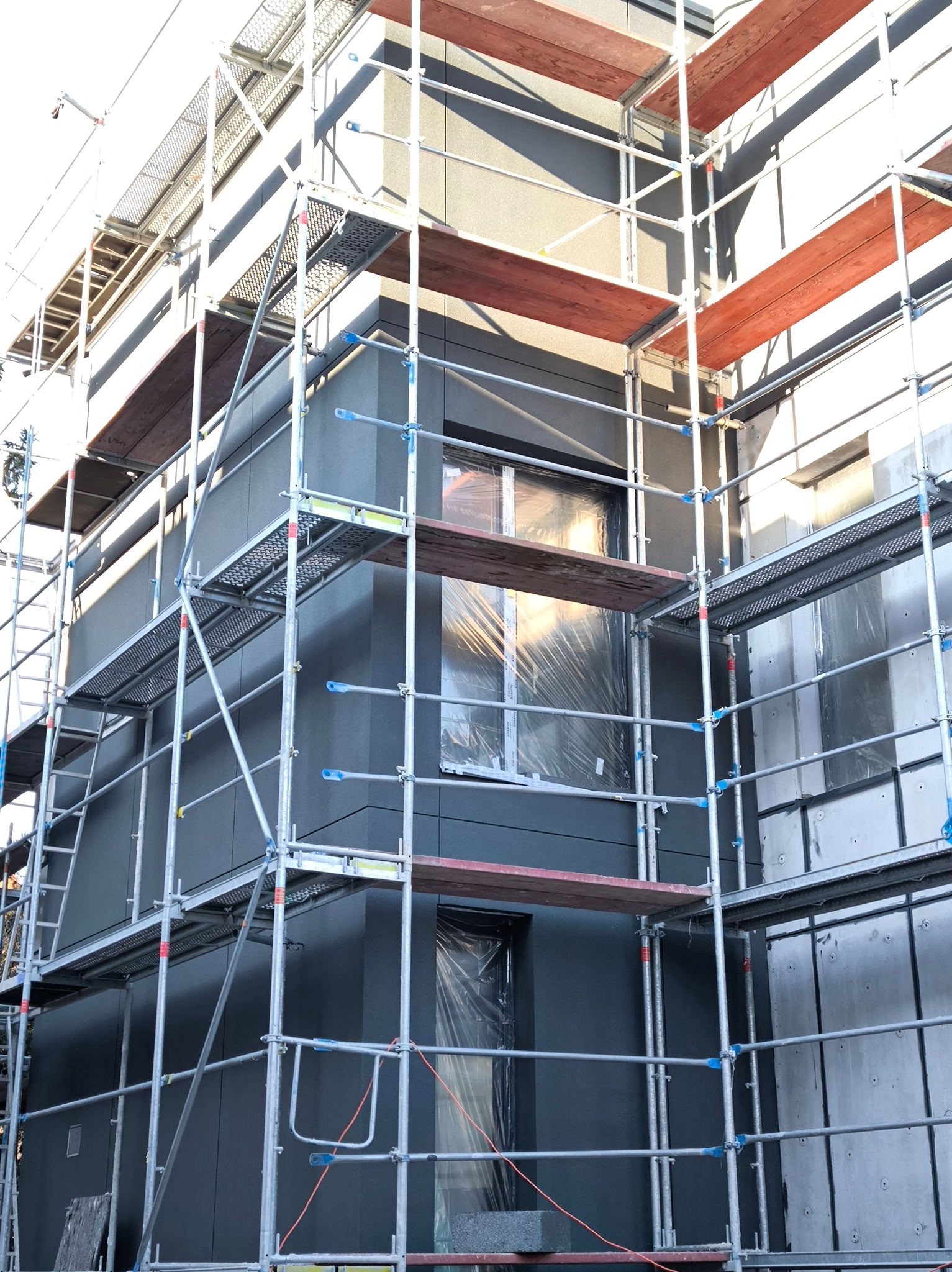 Budynek w trakcie prac elewacyjnych, osłonięte okna folią, widoczne rusztowania. Szara elewacja z panelami, częściowo ocieplona. Ujęcie z dołu, w dziennym świetle.