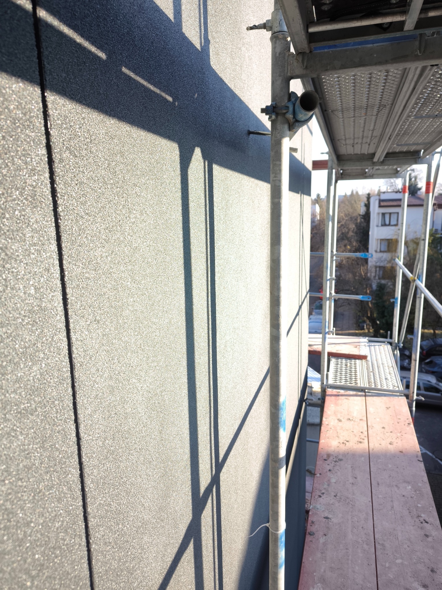 Szara elewacja budynku w trakcie prac wykończeniowych, widoczne rusztowanie i cień. Tekstura ściany z bliska, oświetlona słońcem.