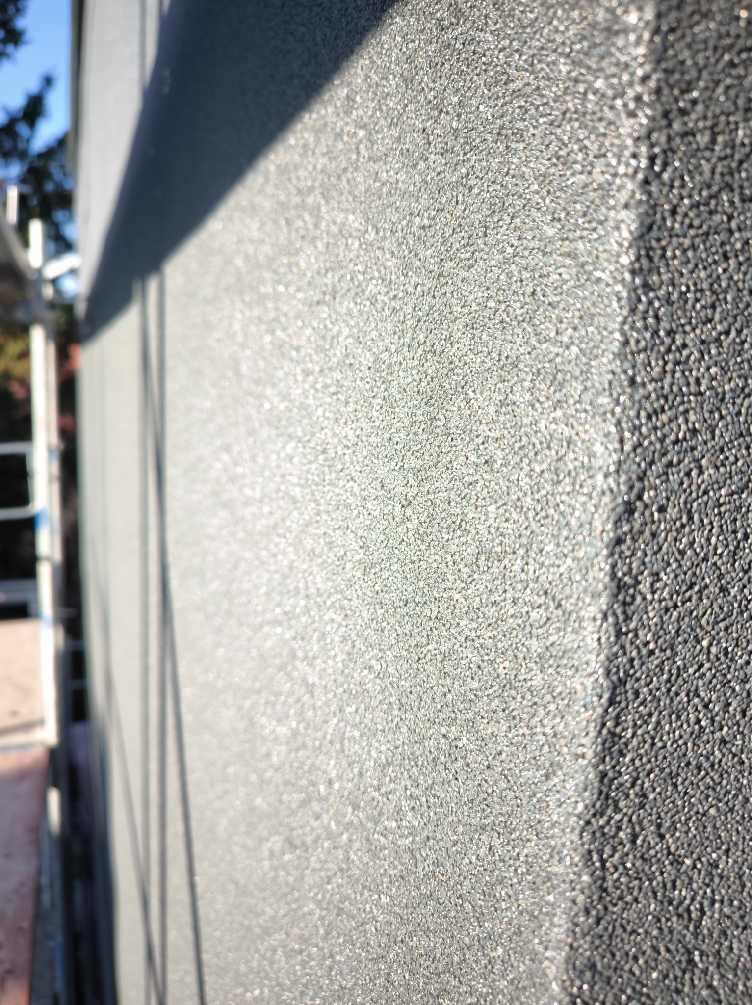 Szara, ziarnista faktura tynku elewacyjnego w zbliżeniu, z widocznym cieniem i fragmentem rusztowania w tle. Delikatne refleksy światła podkreślają strukturę.