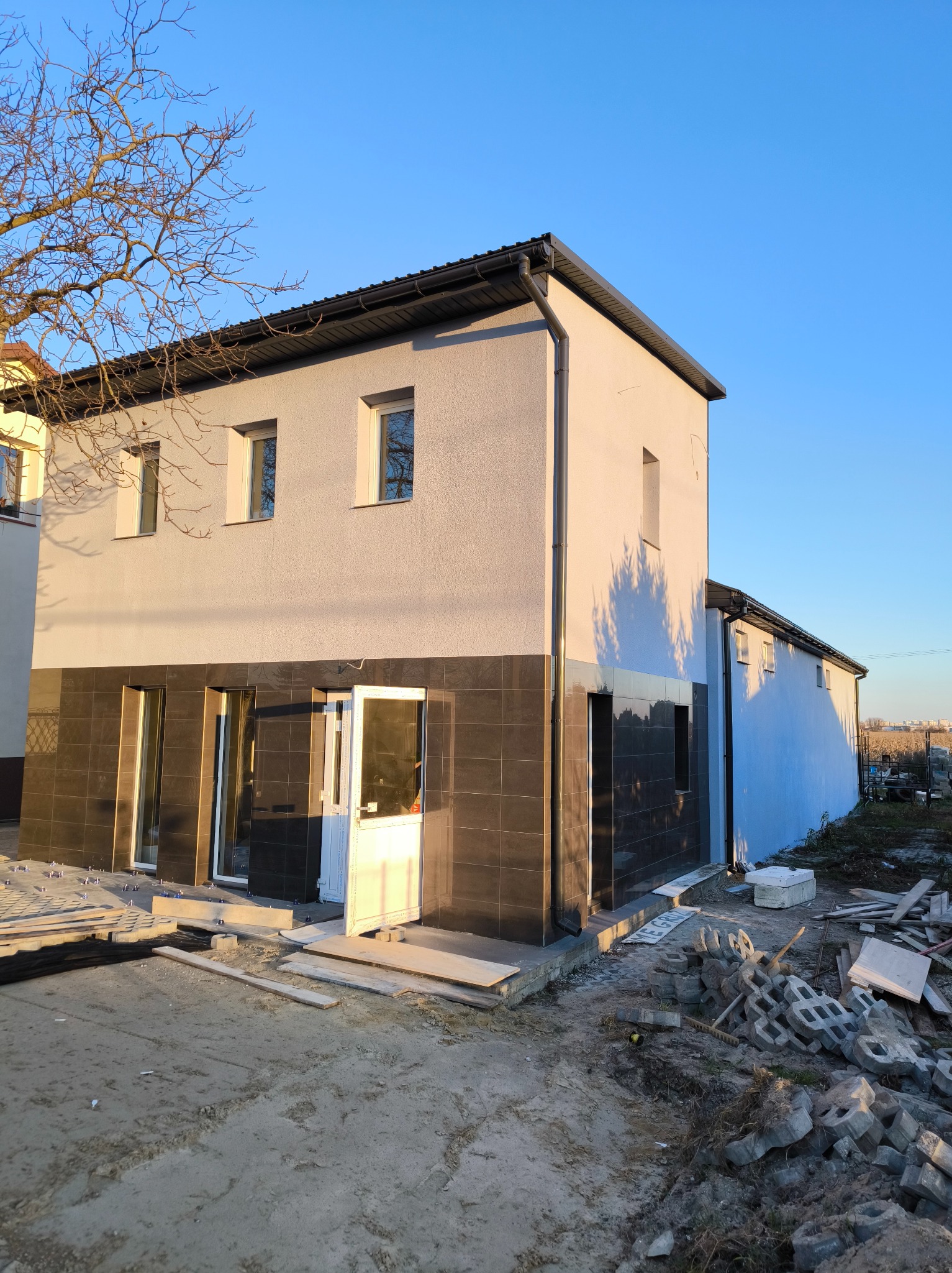 Dwukondygnacyjny budynek z jasną elewacją i ciemną okładziną na dole, w trakcie prac wykończeniowych na zewnątrz, widoczne materiały budowlane i otwarte drzwi.