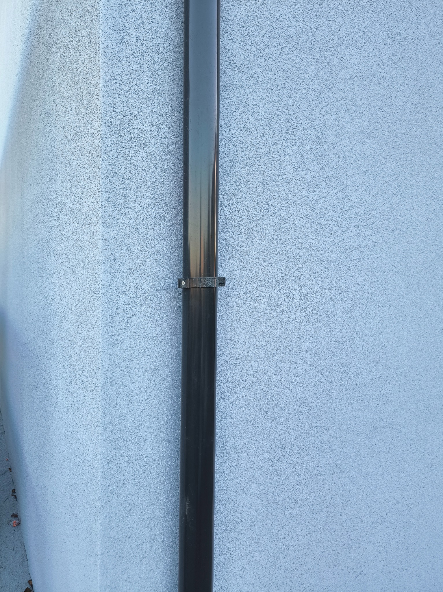 Jasna elewacja budynku z widoczną, pionową rurą spustową, zamocowaną za pomocą metalowych obejm. Tekstura ściany chropowata, równomiernie oświetlona.