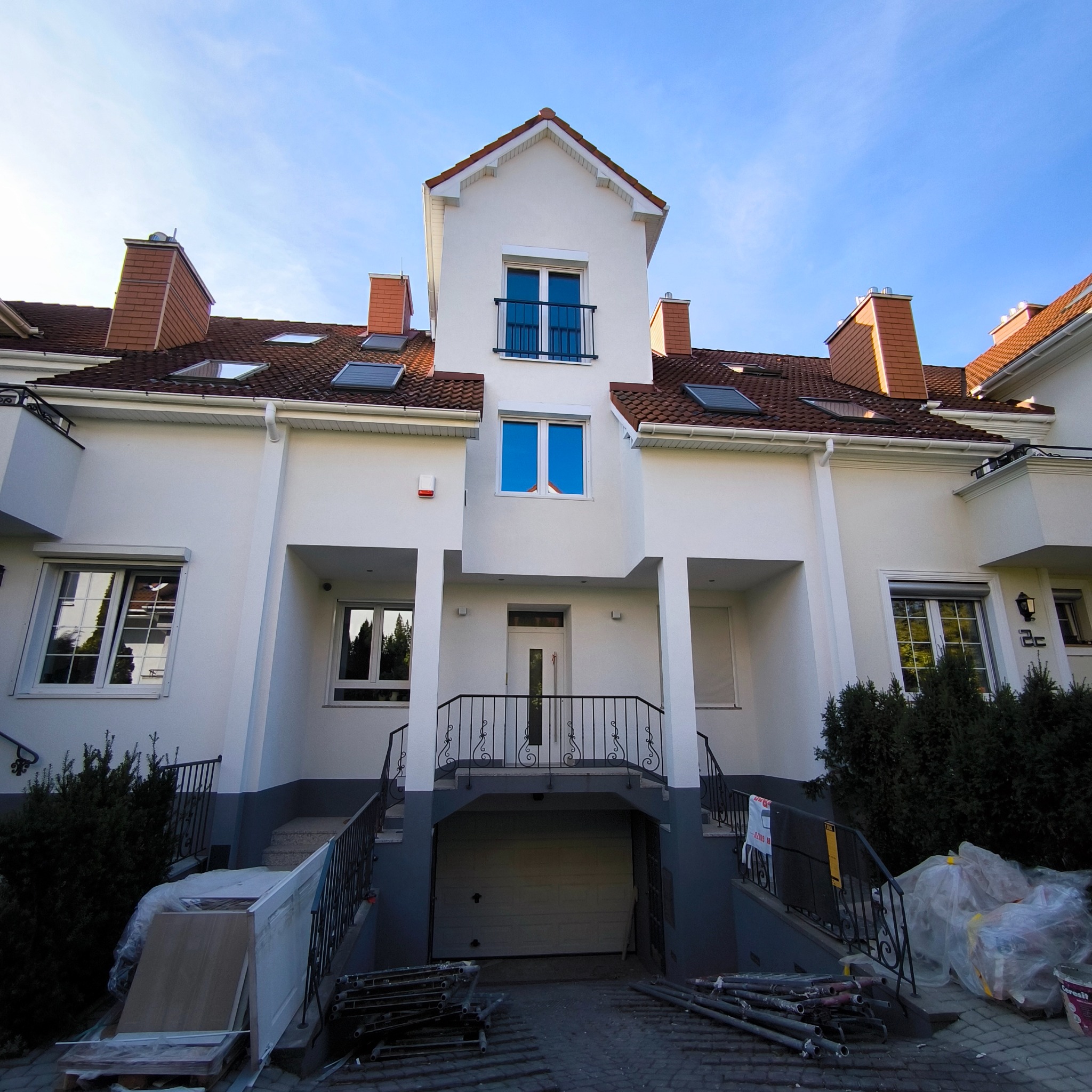 Biała elewacja szeregowca z czerwonym dachem i elementami wykończeniowymi. Na dole widoczne materiały budowlane i rusztowania, sugerujące prace wykończeniowe.