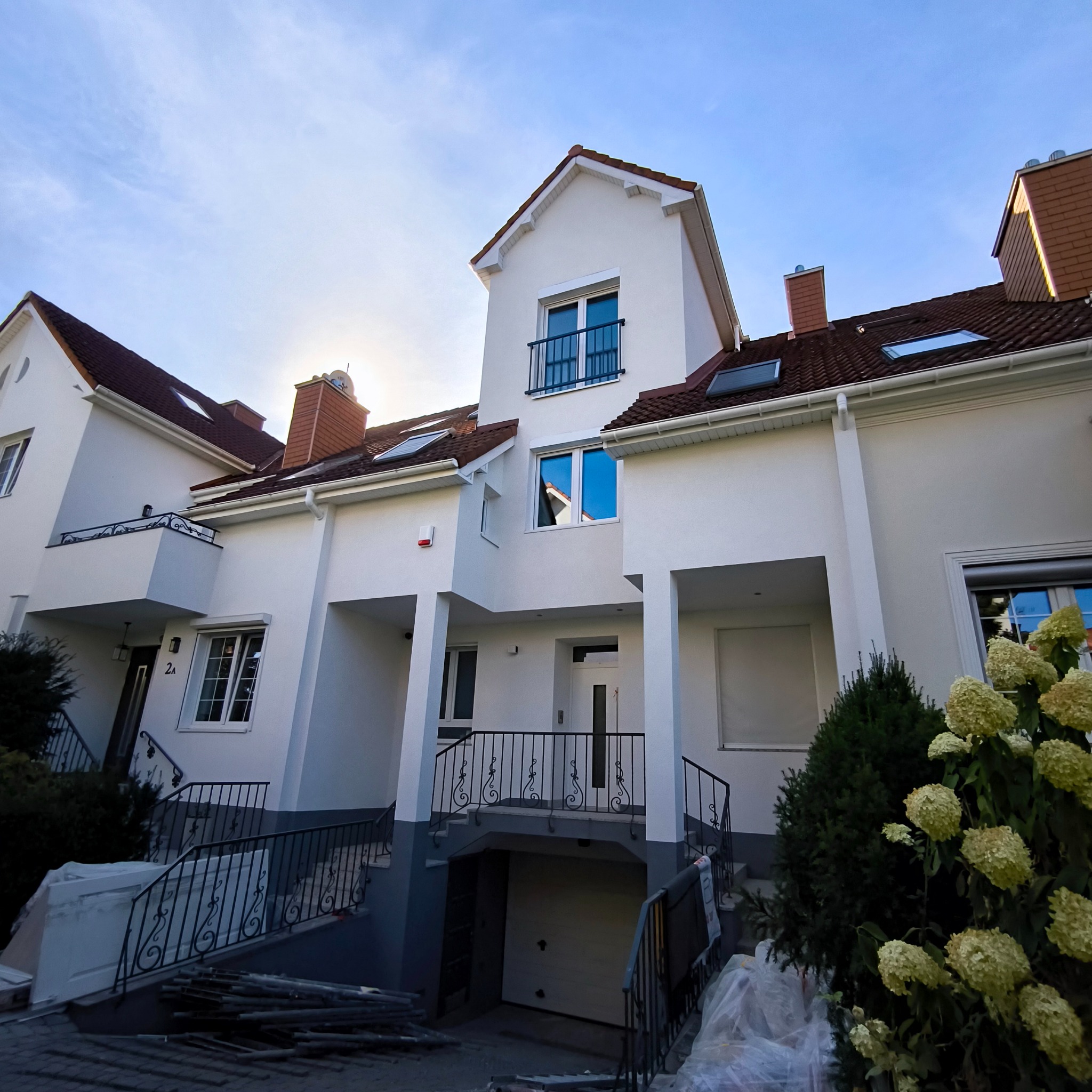 Białe segmenty domów z brązowymi dachami, widoczne z dołu pod kątem. Elementy dekoracyjne i roślinność w otoczeniu. Słońce w tle.