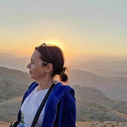 Iwona Sieluk Terapia - Olsztyńskie Centrum Terapii TSR Punkt Zwrotny - Kobieta z włosami spiętymi w kok, w niebieskiej bluzie, patrzy w dal na górzysty krajobraz podczas zachodu słońca, okulary na głowie.