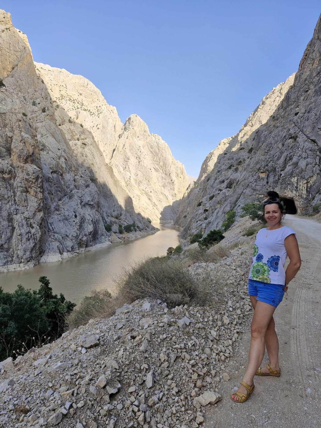 Kobieta w niebieskich spodenkach i białej koszulce z zielono-niebieskim wzorem stoi na kamienistej ścieżce w wąwozie, z rzeką i wysokimi skałami w tle.