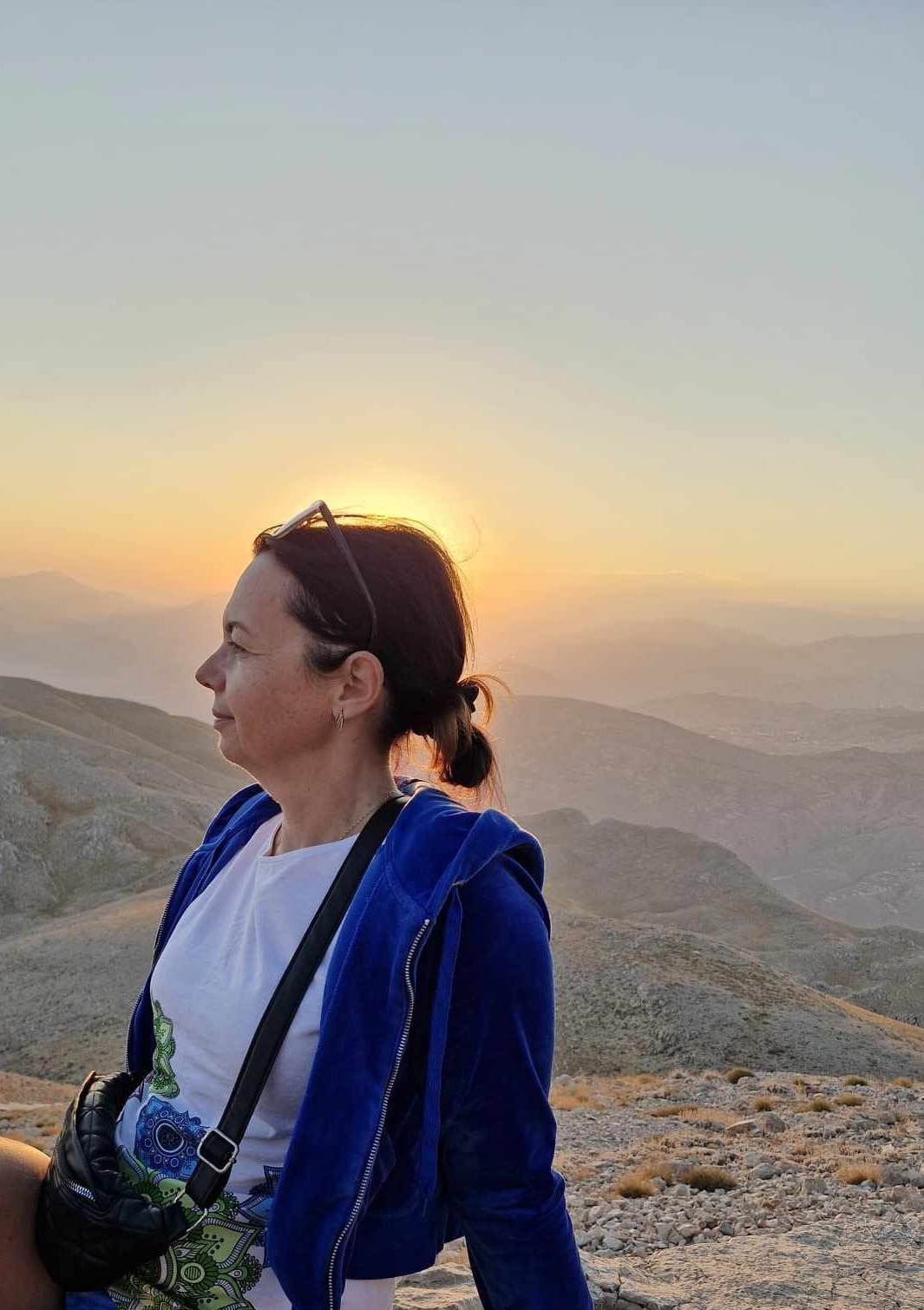 Kobieta z włosami spiętymi w kok, w niebieskiej bluzie, patrzy w dal na górzysty krajobraz podczas zachodu słońca, okulary na głowie.