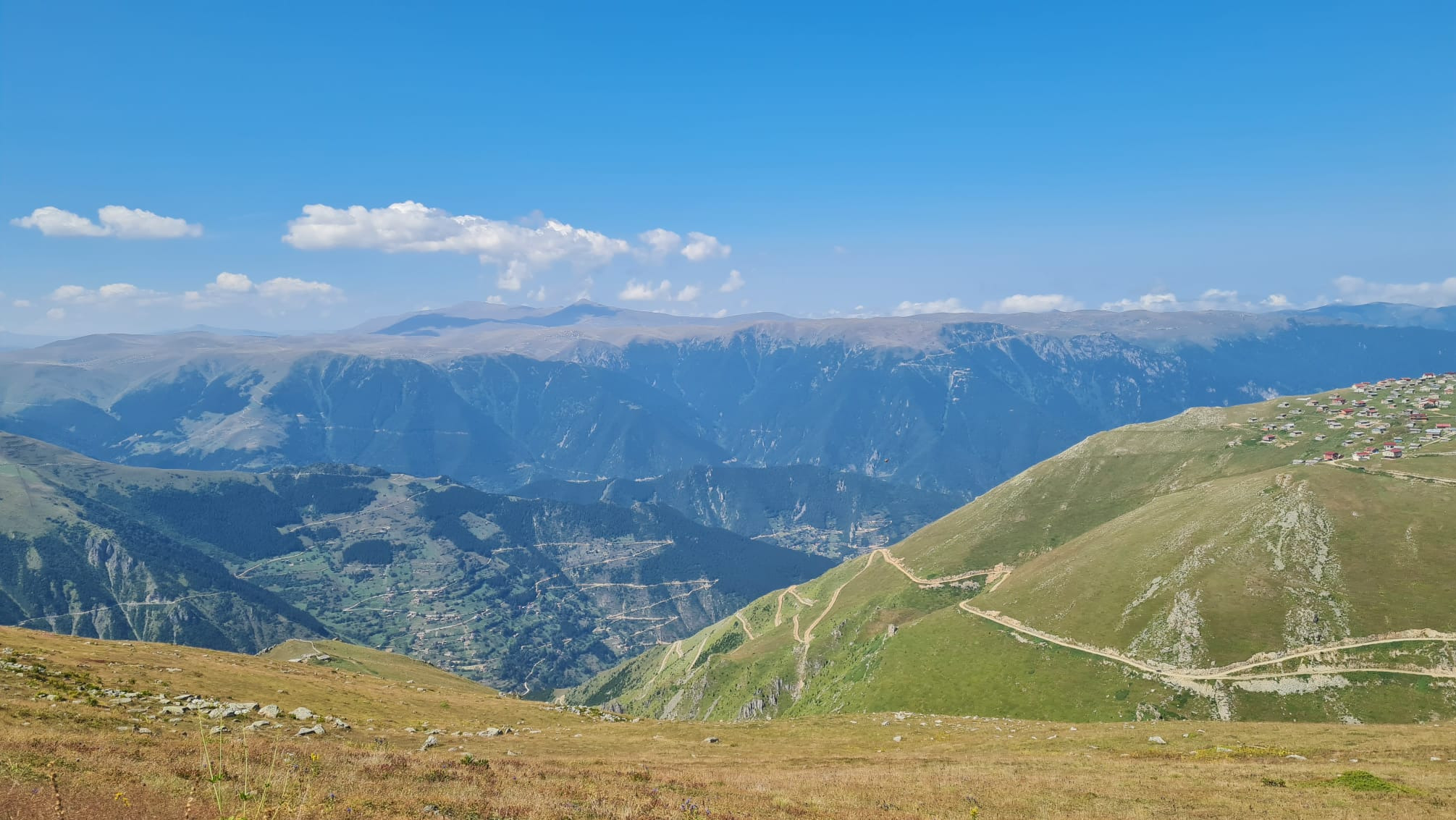 Panoramiczny widok na góry z krętymi ścieżkami i osadą na zboczu, błękitne niebo z białymi obłokami w tle.