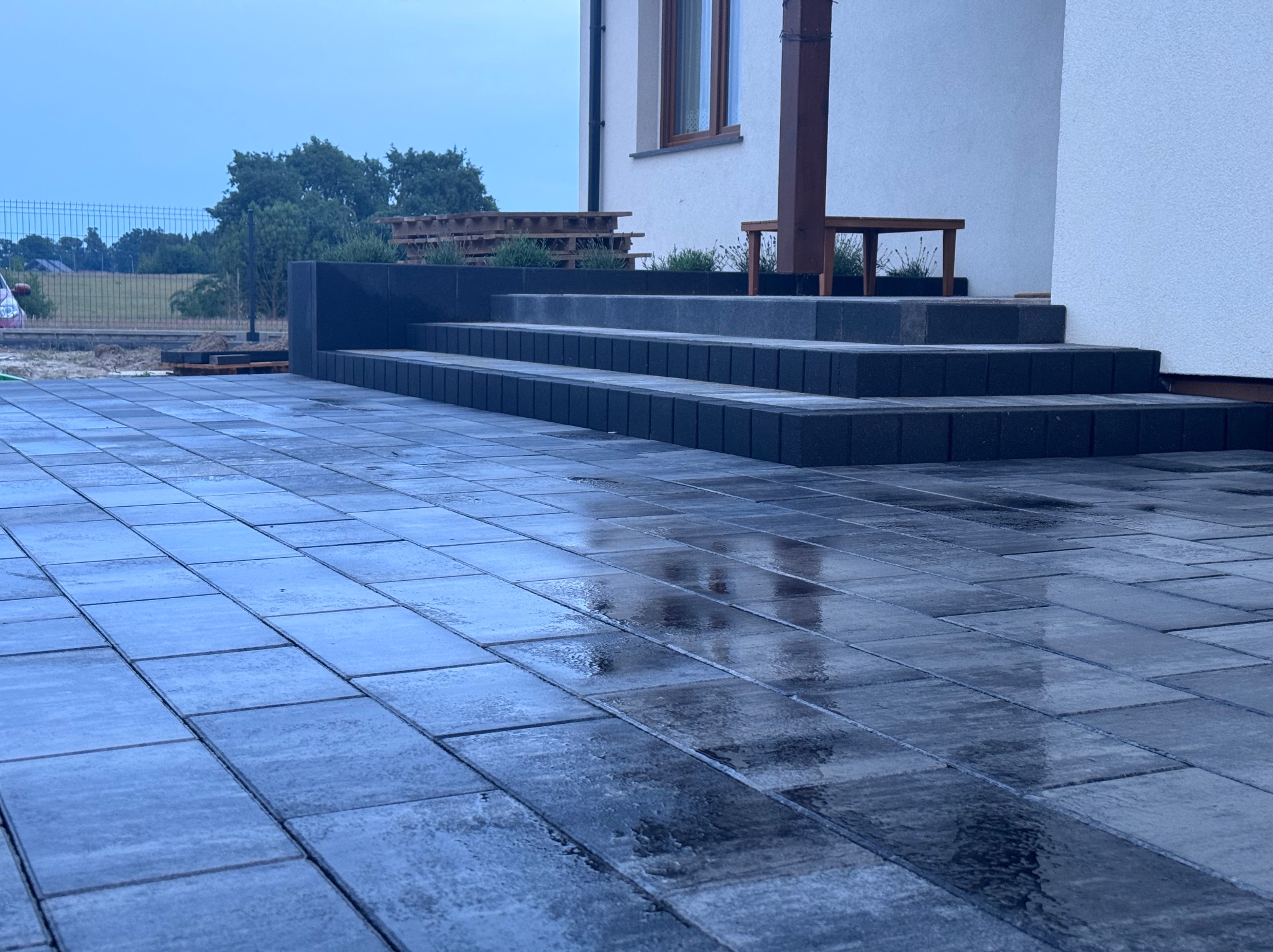 Nowoczesny podjazd z ciemnej kostki brukowej przy domu z schodami wejściowymi, po deszczu. Widoczne odbicia wody na kostce i elementy architektury ogrodowej.