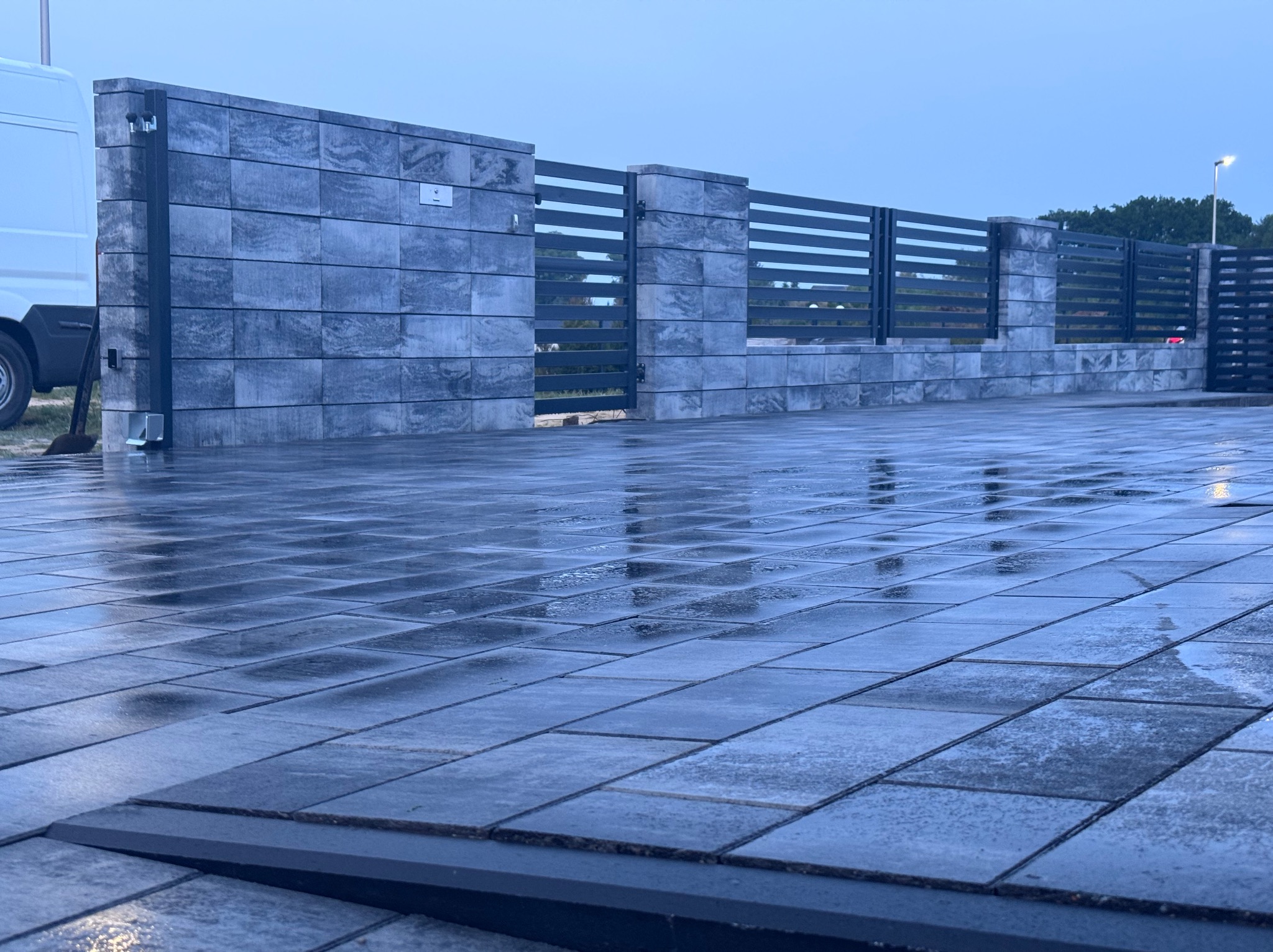 Ułożona, szara kostka brukowa na podjeździe, z fragmentem ogrodzenia z szarych bloczków i czarnych, poziomych przęseł. Powierzchnia mokra, odbija światło.