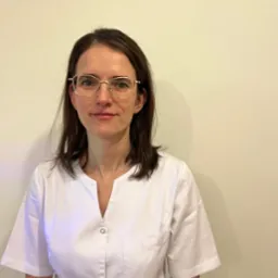 Portret kobiety w białym uniformie medycznym i złotych okularach, stojącej na tle jednolitej, jasnej ściany.