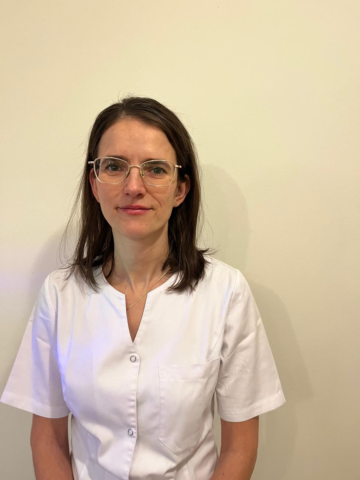 Portret kobiety w białym uniformie medycznym i złotych okularach, stojącej na tle jednolitej, jasnej ściany.