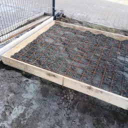Przygotowanie podłoża pod wylewkę betonową: szalunek drewniany, siatka zbrojeniowa i częściowo wylany beton, widoczne także elementy ogrodzenia.