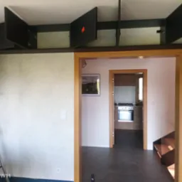 Ściana po nałożeniu gładzi z widoczną drabiną malarską i czarnymi szafkami podsufitowymi z uchylonymi drzwiczkami, w tle widok na kuchnię przez otwarte drzwi.