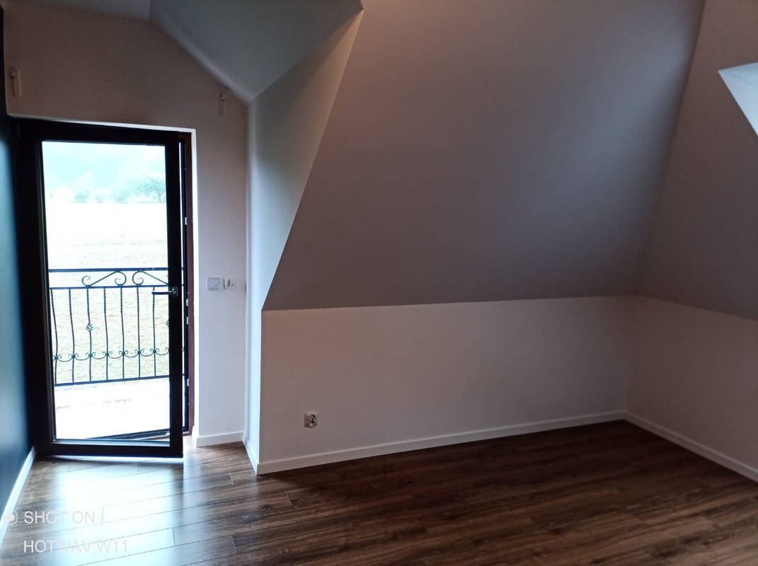 Poddasze z drewnianą podłogą, białe ściany pomalowane do połowy wysokości, widoczne gniazdko elektryczne i drzwi balkonowe z ozdobną balustradą, przez które widać zieleń.