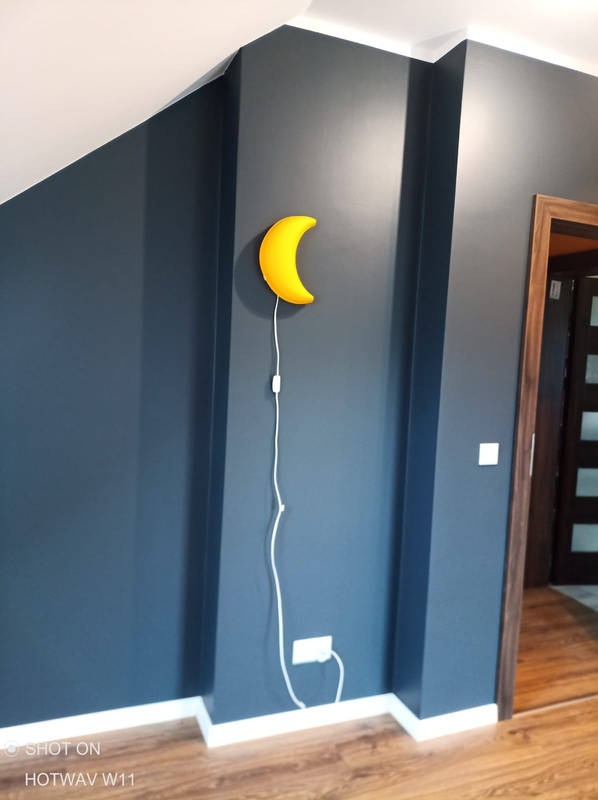 Ściana w pokoju na poddaszu pomalowana na ciemny odcień niebieskiego, z żółtą lampką nocną w kształcie księżyca i widocznym gniazdkiem elektrycznym.