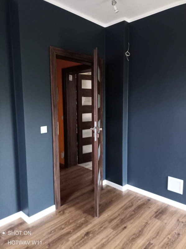 Pomieszczenie po remoncie: ciemnogranatowe ściany, drewniana podłoga, otwarte drzwi z szybami, wystające kable od oświetlenia sufitowego.