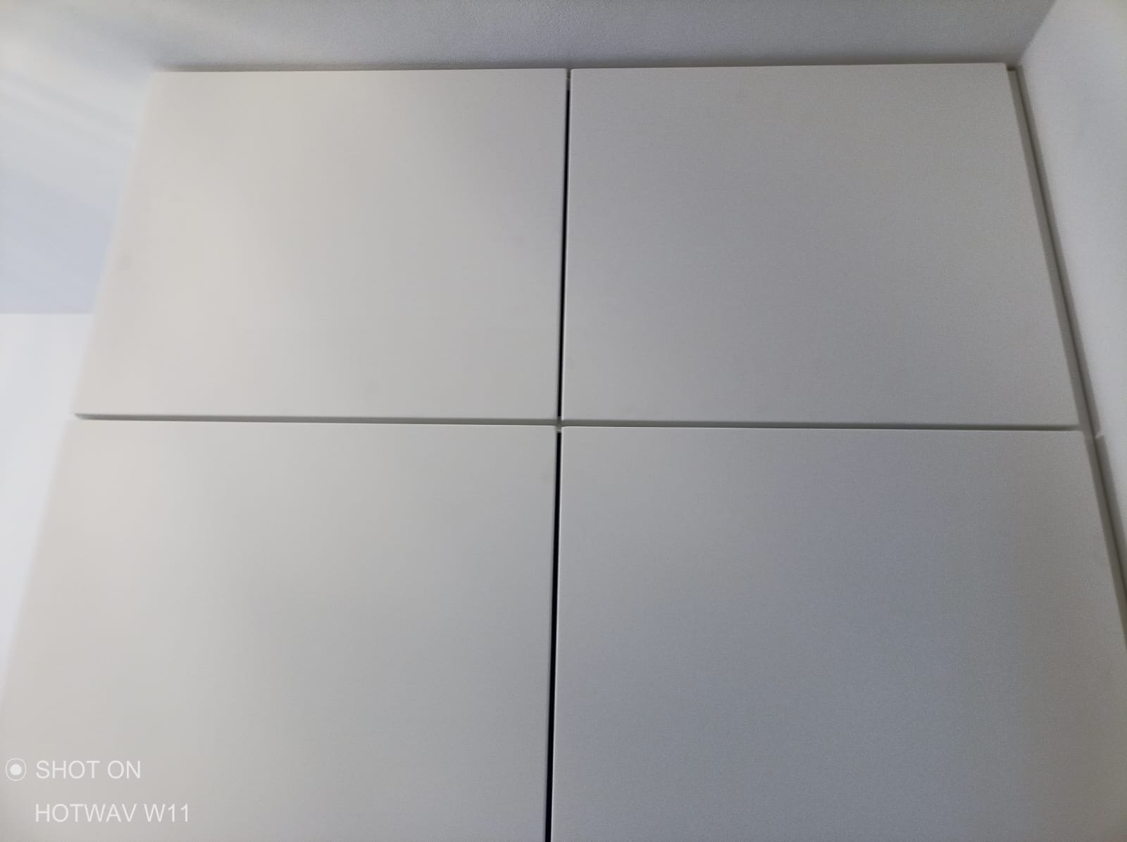 Cztery gładkie, białe, kwadratowe panele, prawdopodobnie fronty szafek, z delikatnymi szczelinami między nimi, sfotografowane pod kątem.