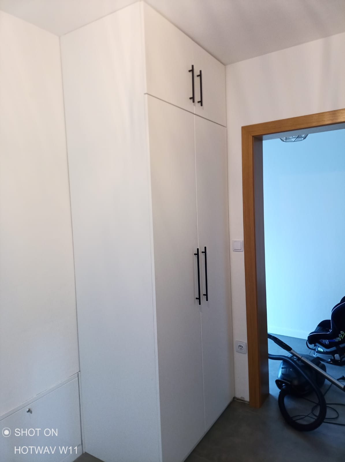 Biała, wysoka szafa wnękowa z czarnymi, pionowymi uchwytami, widoczna przy drewnianej ościeżnicy drzwi, z odkurzaczem i fotelikiem dziecięcym w tle.