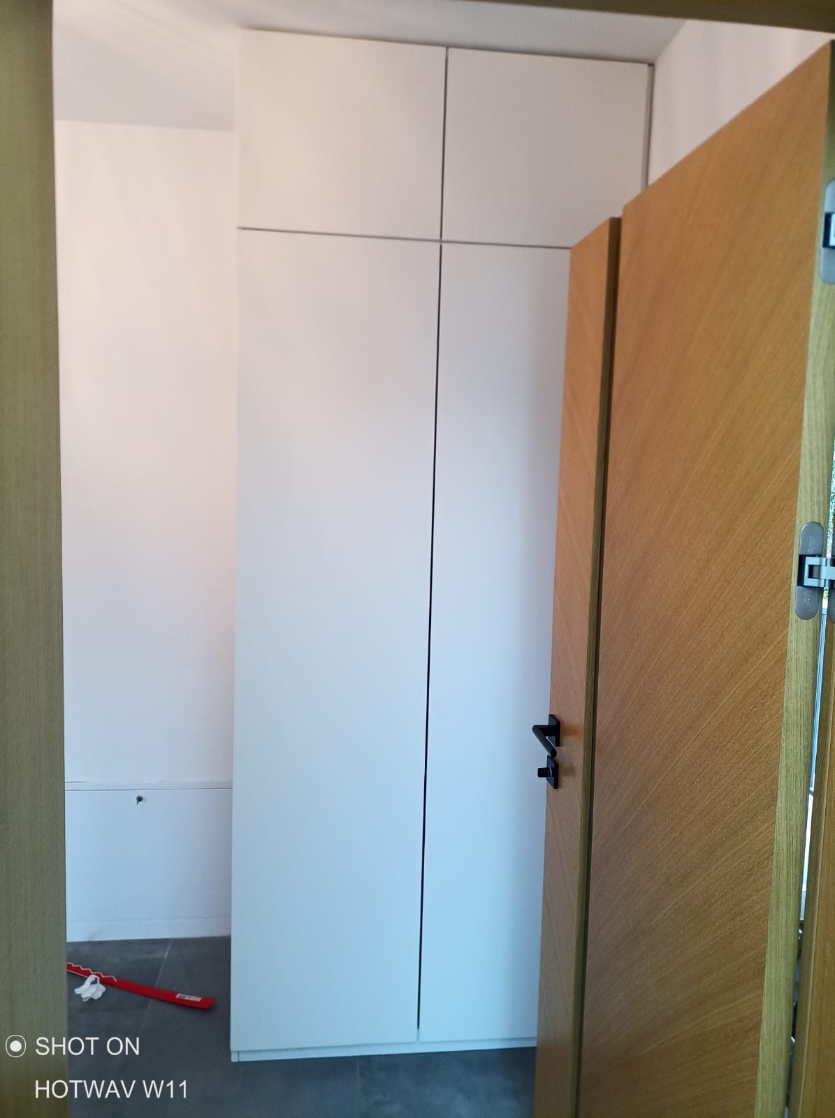 Biała zabudowa szafy wnękowej z drewnianymi drzwiami po prawej stronie, widok z perspektywy wnętrza, na podłodze widoczny czerwony element.