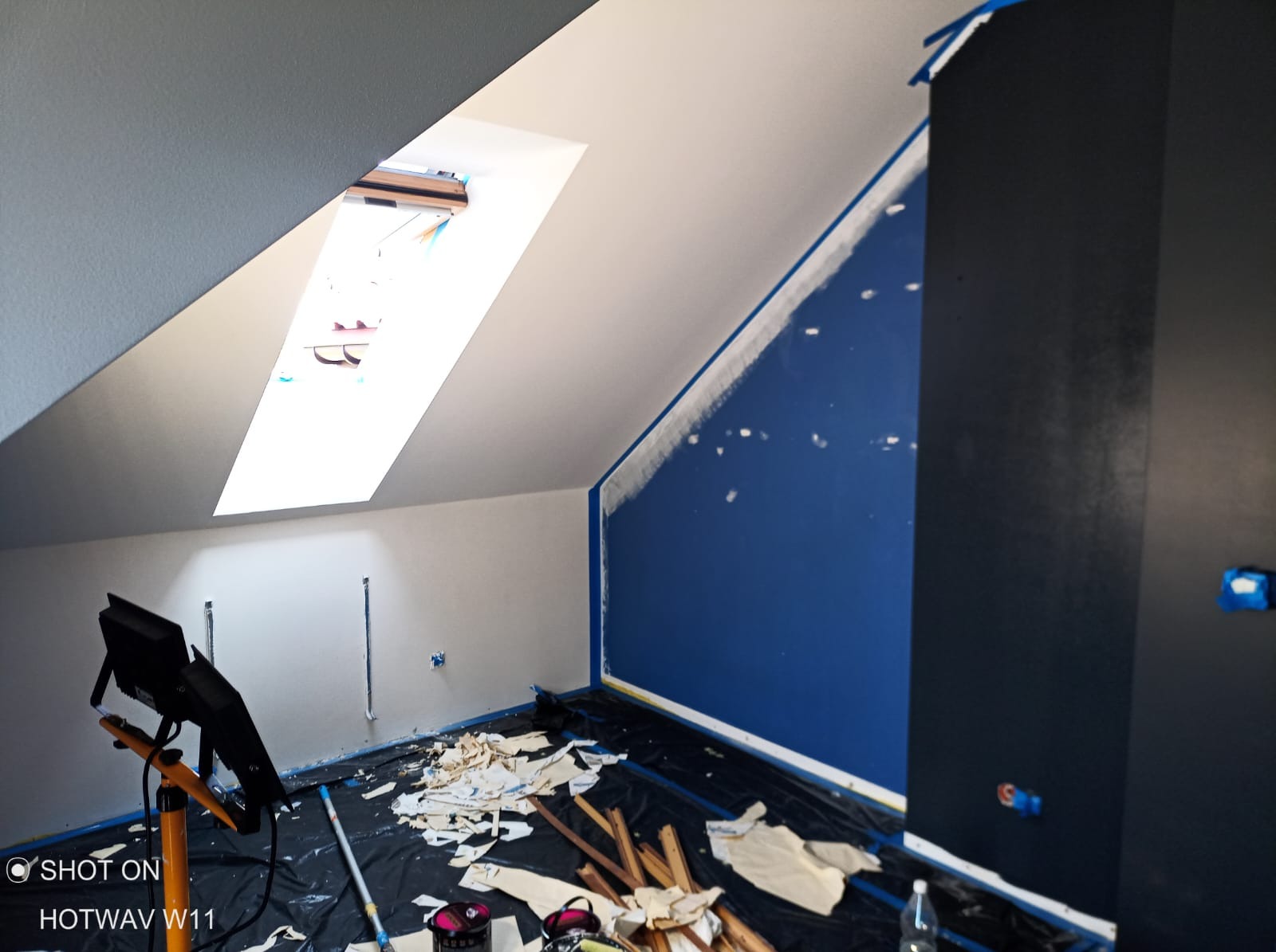 Poddasze w trakcie prac wykończeniowych: ściany częściowo pomalowane na biało i granatowo, zabezpieczone taśmą malarską, podłoga przykryta folią z resztkami tapet, widoczna lampa halogenowa...