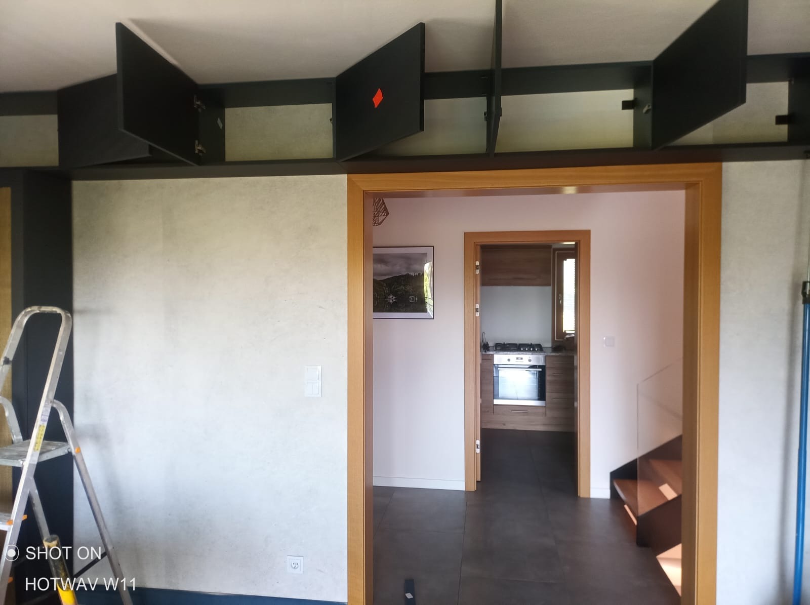 Ściana po nałożeniu gładzi z widoczną drabiną malarską i czarnymi szafkami podsufitowymi z uchylonymi drzwiczkami, w tle widok na kuchnię przez otwarte drzwi.