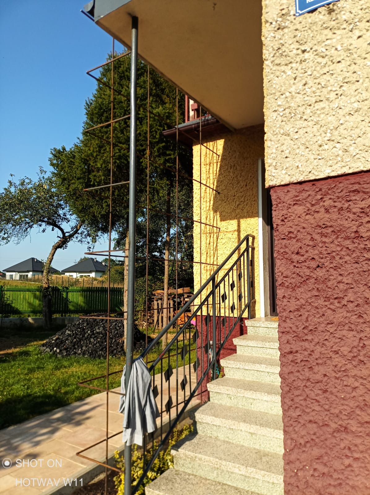 Zewnętrzne schody wejściowe z szarego granitu prowadzące do domu z elewacją w odcieniach beżu i bordo, widoczna metalowa balustrada, na zewnątrz drzewa i trawnik.