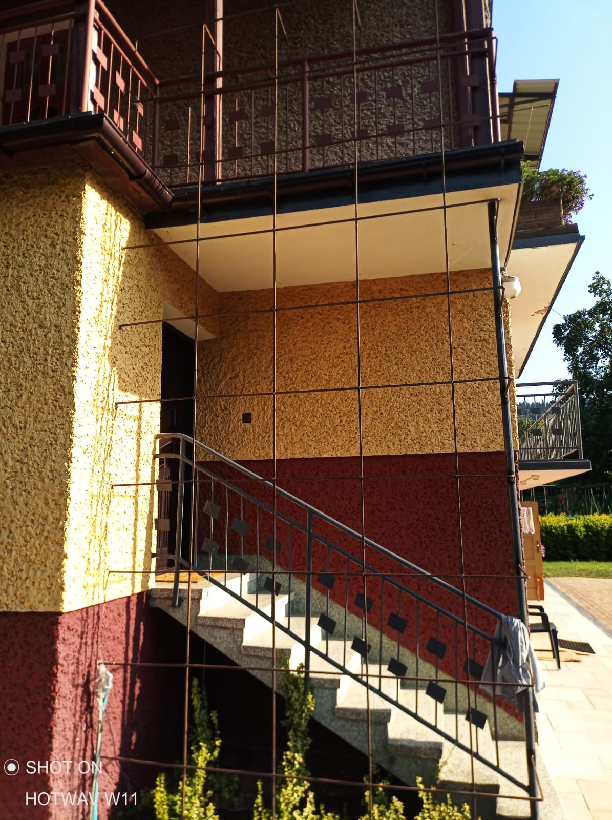 Elewacja budynku z widocznymi schodami wejściowymi, z nałożoną siatką ochronną, ściana w odcieniach żółci, pomarańczy i czerwieni, z fakturą baranka.