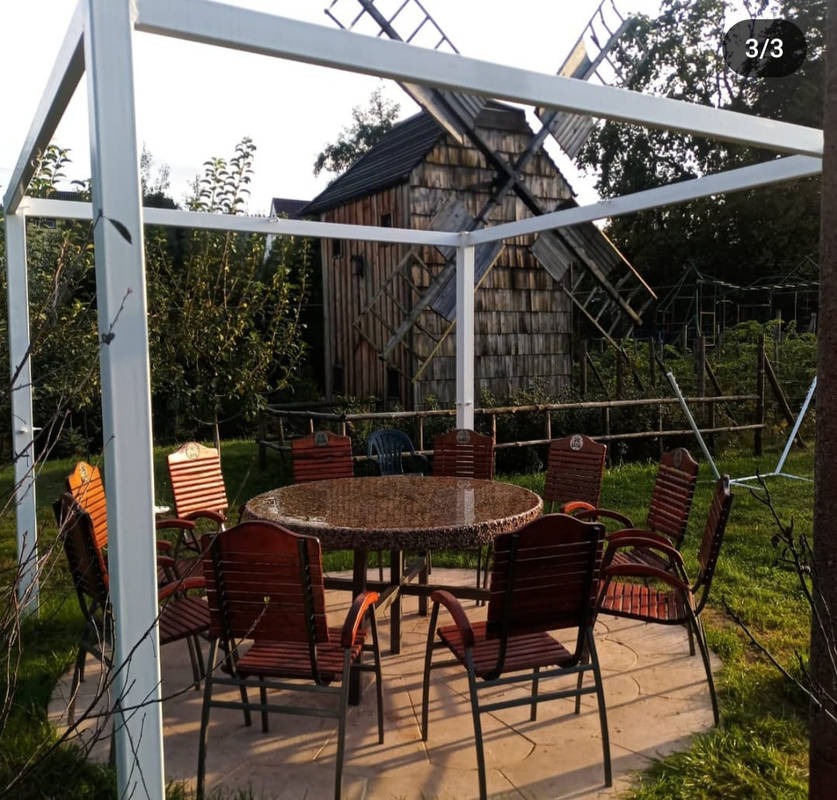 Ogród z zadaszeniem pergolowym, okrągłym stołem i krzesłami na tle drewnianego wiatraka.