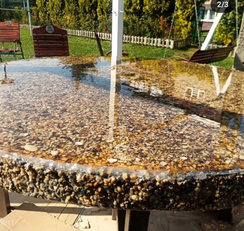 Okrągły stół ogrodowy z blatem z żywicy epoksydowej wypełnionym drobnymi kamykami, widoczne odbicie otoczenia w błyszczącej powierzchni, w tle trawnik, drzewa i huśtawka.