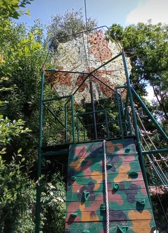 Zewnętrzna konstrukcja wspinaczkowa z elementami kamuflażu i siatką maskującą na szczycie, otoczona zielenią.
