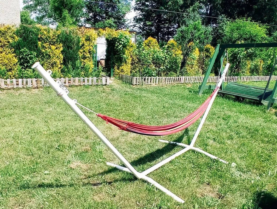 Czerwono-niebieski hamak zawieszony na białym metalowym stelażu, ustawiony na trawniku w ogrodzie z drewnianym płotem i zielonymi krzewami w tle.