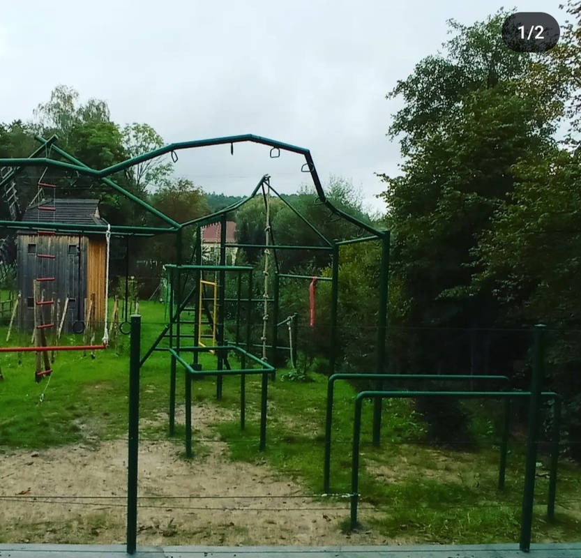 Zielony plac zabaw na trawiastym terenie z drewnianą konstrukcją w tle, widok w pochmurny dzień.