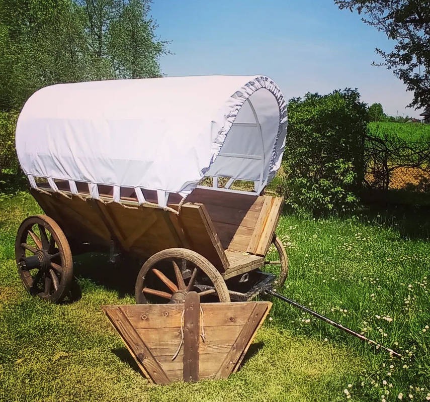 Rustykalny, drewniany wóz z białym płóciennym dachem, stojący na trawiastym terenie z widocznym fragmentem drewnianej skrzyni przed nim, na tle zieleni drzew i krzewów.